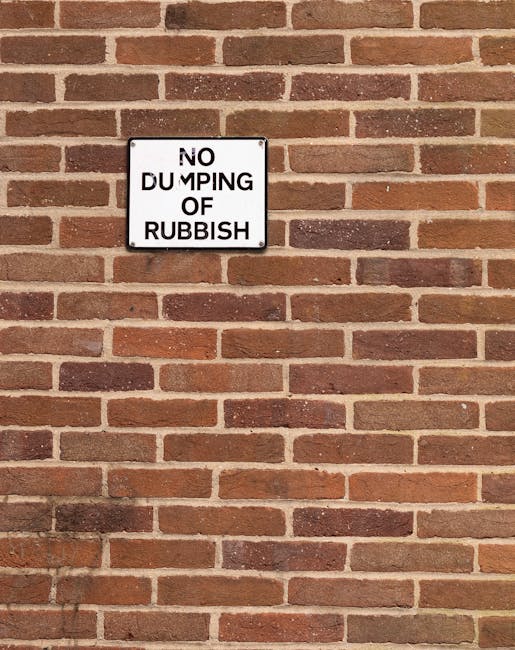 A close-up photograph of a brick wall made of reddish-brown squared bricks with variations in shade, arranged in a horizontal running bond pattern. Attached to the wall is a small rectangular metal sign with a white background and black text that reads 'No Dumping of Rubbish,' secured with visible bolts at each corner. The bricks have a rough texture with some mortar joints showing signs of weathering and slight discoloration. The wall is evenly lit with soft, natural lighting, and the sign is positioned centrally within the image, emphasizing the message against the background of the brick surface. The scene reflects an outdoor location where waste disposal is regulated and monitored, consistent with rubbish removal and property management contexts often handled by services like Waste Clearance Catford.