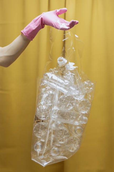 A person wearing a pink rubber glove is holding a large, transparent plastic bag filled with crumpled and flattened clear plastic bottles and containers. The background features a yellow curtain or wall, providing a solid, vibrant backdrop. The plastic items inside the bag are of various shapes and sizes, with some caps and labels visible, indicating they are recyclable materials. The scene appears to be indoors, possibly in a waste sorting or recycling context. The image emphasizes the importance of proper waste segregation and recycling practices, which are part of effective rubbish removal services offered by Waste Clearance Catford. The clear plastic waste highlights the need for responsible disposal and could relate to private disposal options for recyclable materials, in contrast to municipal waste collection. The detailed textures of the plastic bottles, combined with the soft lighting, serve to underscore environmentally conscious waste handling.