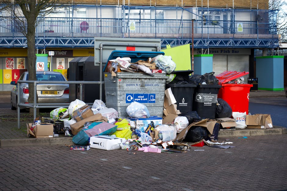A large pile of discarded rubbish and waste materials is accumulated on a section of paved pavement in an urban setting, including a variety of cardboard boxes, plastic bags filled with trash, crumpled paper, and miscellaneous packaging. The waste is scattered in front of a metal recycling bin labeled for mixed paper and cardboard, which is partially overflowing with torn paper, cardboard, and other refuse. Nearby, there are several black and red waste bins, some with closed lids and others open, revealing more trash. To the left, a parked silver station wagon is visible with a license plate. In the background, there is a commercial building under renovation or construction, covered in blue scaffolding with protective netting, and a storefront with signage indicating local shops beneath. The scene is illuminated by natural daylight, and the overall environment appears to be a typical urban waste collection point potentially serviced by private waste management providers such as Waste Clearance Catford, aligning with alternative waste disposal methods outside local authority collection.