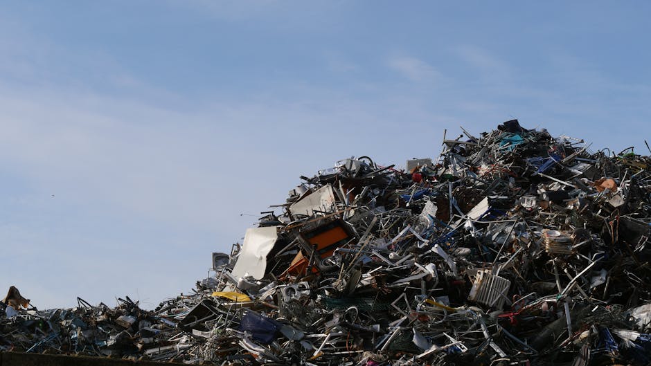 A large open heap of mixed metal and electronic waste is seen outdoors against a clear blue sky. The pile contains various discarded appliances, metal frames, wires, and broken electronic devices, with some items showing rust or damage. The materials have different textures, from smooth painted panels to rough, jagged metal edges. The waste is piled on a concrete ground, with no visible containment or covering, indicating an unorganized disposal site. The environmental context suggests a site dedicated to scrap or waste accumulation, possibly for recycling or collection services like those provided by Waste Clearance Catford. The bright daylight illuminates the scene, highlighting the complex arrangement and varied materials within the waste heap, reflecting the nature of independent or alternative waste handling methods outside standard rubbish collection channels.