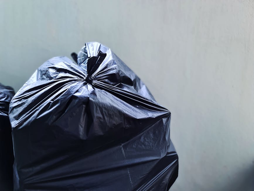 Close-up of a large black plastic rubbish bag tightly sealed at the top, placed against a plain, light-colored wall. The bag appears to be made of glossy, durable material with visible creases and folds, indicating it is filled with waste debris. It is positioned slightly to the left of the frame, with part of another similar bag partially visible on the far left side. The plain background emphasizes the focus on the rubbish bag, which is likely part of a waste collection or disposal process. The scene suggests an environment where waste is collected for removal, consistent with private rubbish clearance services, such as those provided by Waste Clearance Catford. The lighting is neutral and soft, casting minimal shadows, and further highlights the glossy finish of the plastic. This image can be associated with on-site waste management or alternative waste handling methods outside of standard local authority collection, aligning with professional rubbish removal activities.