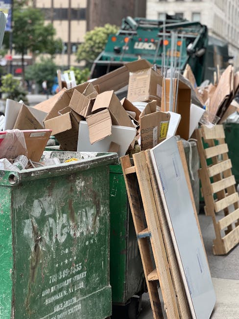 A large outdoor area shows a significant accumulation of waste, primarily consisting of stacked boxes of fresh fruit and vegetable produce, identifiable by their colorful packaging and printed labels. The boxes, made of cardboard and featuring a glossy finish, are arranged in multiple piles, some leaning slightly, with visible stacking patterns on a paved surface. Surrounding the produce boxes are various black plastic crates, some stacked on top of each other, along with cardboard boxes and other packaging materials. To the right, metal wire cages are filled with assorted cardboard boxes and packaging, some partially open revealing contents within, while others are sealed with packing tape. In the foreground, several closed wheelie bins with green and red lids are positioned on the pavement, with some partially overlapping. The background includes a glimpse of a garden or outdoor landscape, with trees, grass, and distant houses, illuminated by natural daylight which provides clear visibility of all objects. The scene suggests a private waste clearance operation by Waste Clearance Catford, involving the collection of mixed waste items typical of bulk rubbish removal or alternative waste handling services, captured in a professional and factual manner.