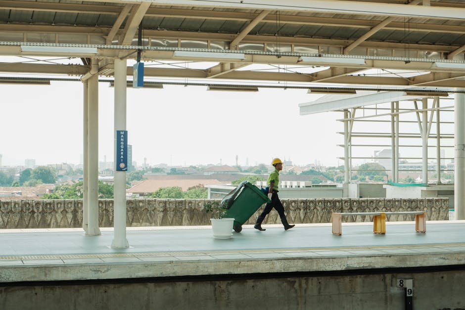 A person wearing a yellow safety helmet, green high-visibility vest, and dark trousers is walking along an elevated outdoor platform, pulling a large green wheeled rubbish bin behind them. The bin appears to be made of durable plastic with a lid secured on top. They are walking next to a white plastic bucket positioned on the platform surface. The platform itself is constructed from sturdy concrete and features a canopy overhead with a metal frame, providing partial shade. To the right, there are three small, orange and white plastic barriers or seating stools arranged along the edge of the platform. In the background, a low decorative stone wall separates the platform from an expansive view of distant rooftops and trees under a bright, overcast sky. The scene suggests an organized urban waste collection or on-site clearance activity, indicative of private waste handling services such as those provided by Waste Clearance Catford, supporting alternative rubbish removal options outside standard municipal collection. The environment appears clean, with no visible litter or clutter, emphasizing professional waste management practices.