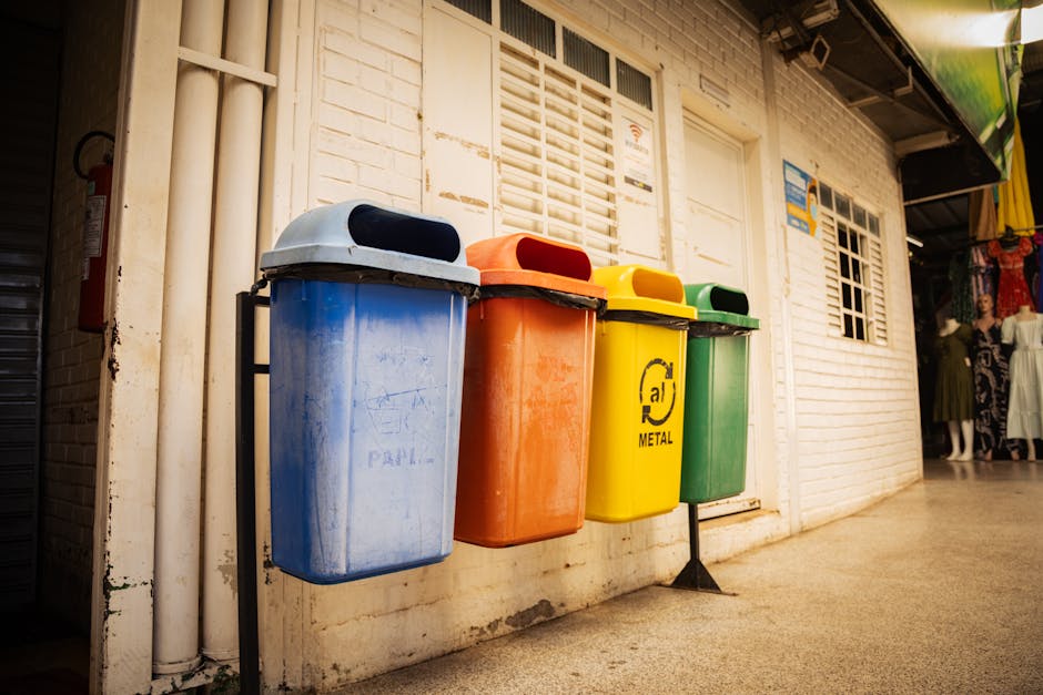 Four waste bins lined up against a white brick wall outside a building, each with different coloured lids: blue, orange, yellow, and green. The blue bin on the left has a rounded grey lid, possibly for general waste, showing some dirt and minor graffiti markings on its surface. Adjacent to it, the orange bin features a darker rubber lid and appears to be for non-recyclable waste, with visible scuff marks. Next to the orange bin, the yellow bin is marked with a black recycling symbol and the word 'METAL,' indicating its purpose for metal recyclables; its surface looks slightly worn but clean. Finally, the green bin on the far right has a matching green lid, likely designated for glass or other recyclable materials; it sits directly on the paved ground without a visible stand. The bins are positioned in a narrow outdoor corridor, possibly near a commercial or residential space, with environmental lighting suggesting daylight. Waste Clearance Catford might provide solutions for private waste handling or non-local-authority rubbish disposal, as indicated by the presence of these designated waste containers outside the premises.