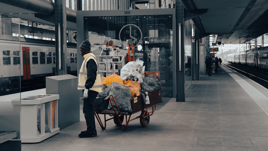 A person wearing a yellow safety helmet, green high-visibility vest, and dark trousers is walking along an elevated outdoor platform, pulling a large green wheeled rubbish bin behind them. The bin appears to be made of durable plastic with a lid secured on top. They are walking next to a white plastic bucket positioned on the platform surface. The platform itself is constructed from sturdy concrete and features a canopy overhead with a metal frame, providing partial shade. To the right, there are three small, orange and white plastic barriers or seating stools arranged along the edge of the platform. In the background, a low decorative stone wall separates the platform from an expansive view of distant rooftops and trees under a bright, overcast sky. The scene suggests an organized urban waste collection or on-site clearance activity, indicative of private waste handling services such as those provided by Waste Clearance Catford, supporting alternative rubbish removal options outside standard municipal collection. The environment appears clean, with no visible litter or clutter, emphasizing professional waste management practices.