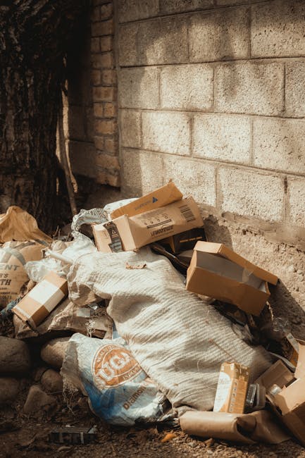 A pile of mixed waste materials and packaging sits outdoors against a beige brick wall and a tree trunk on the left side. The debris includes cardboard boxes, some flattened and others partially open, along with white and brown paper bags and crumpled packaging. The cardboard boxes vary in size and color, with some showing printed labels, and are stacked irregularly, leaning against each other and the wall. A large fabric sack or bag is part of the pile, partially covered by the boxes. The ground beneath the rubbish appears uneven and rocky, with small stones scattered around. The scene has natural lighting, with shadows cast by the objects and the tree trunk, creating a utilitarian environment typical of an outdoor waste collection area or a site awaiting rubbish removal. This visual context aligns with independent waste disposal or rubbish clearance services provided by Waste Clearance Catford, highlighting the need for timely rubbish removal, particularly in private or non-communal settings.