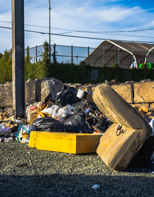 An outdoor scene featuring a pile of discarded rubbish on a gravel surface, including black plastic garbage bags, a yellow plastic container, an old beige car seat, and various other waste items. The rubbish is positioned in front of a low stone wall made of uneven, weathered stones, with some green foliage and bushes visible behind it. In the background, there is a dark metal fence with vertical bars and a large tent-like structure with a curved roof, supported by metal frames, partially visible under a bright, clear blue sky with a few scattered clouds. Overhead, power lines stretch across the scene, attached to wooden utility poles. The lighting indicates daytime with natural sunlight casting shadows on the rubbish, emphasizing the textures of the different materials. The scene suggests a private or independent rubbish collection or waste removal effort, possibly involving a service like Waste Clearance Catford, that is aimed at clearing accumulated waste outside a residential or commercial property.